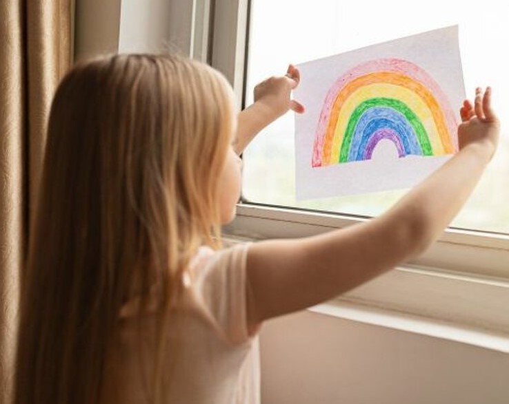 Foto von einem Mädchen welches einen selbstgemalten Regenbogen an ein Fenster hält und dabei lacht